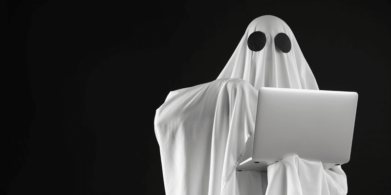 A figure draped in a white sheet with eye holes, ghost-like, stands against black, holding an open laptop—symbolizing ghost users.